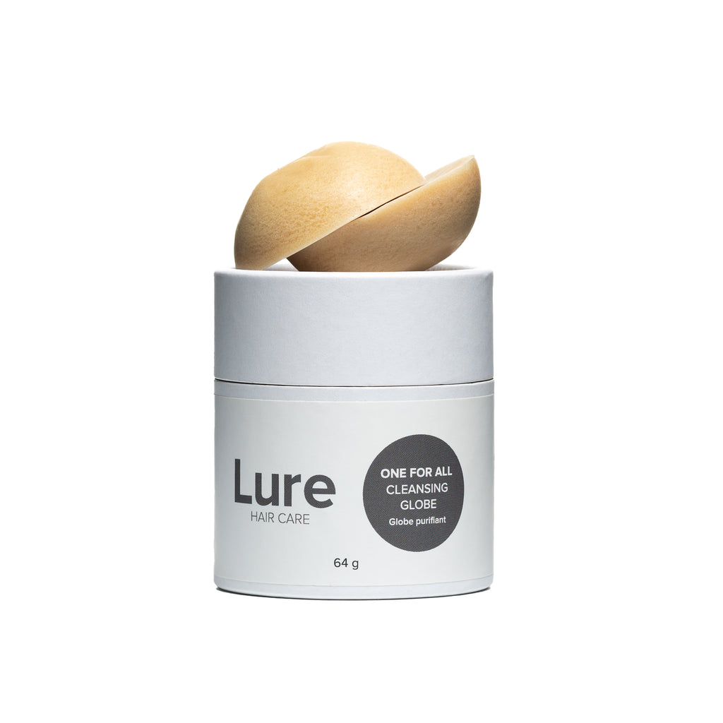 White paper tube with grey font and logo containing Lure hair care's One For All Cleansing Globe.  On top of the tube are two hemispheres of the solid cream cleanser.